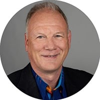 Porträt eines lächelnden Mannes im Anzug und buntem Hemd, grauer Hintergrund.