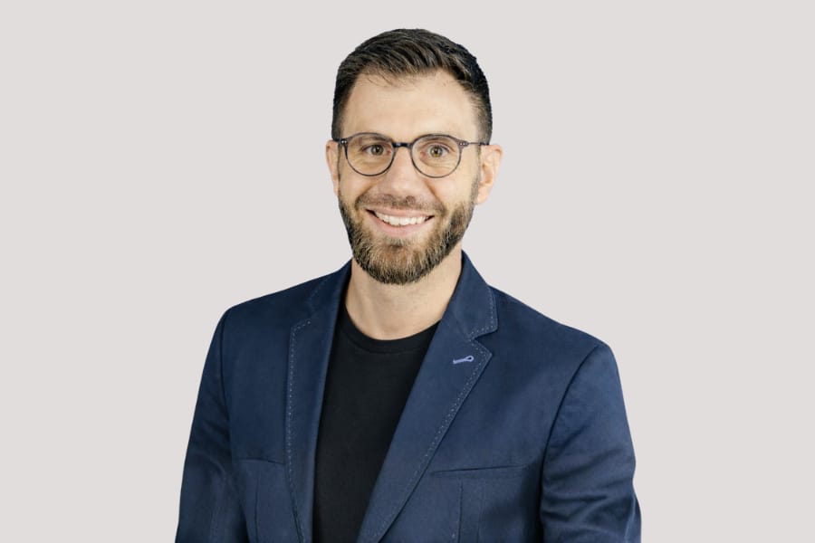 Mann mit Brille in blauem Blazer, lächelnd vor grauem Hintergrund.