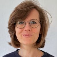 Person mit Brille und braunen Haaren vor neutralem Hintergrund.