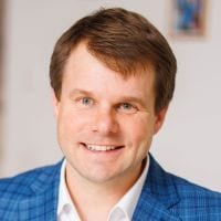 Lächelnder Mann in blauem Anzug vor unscharfem Hintergrund.