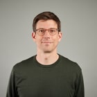 Porträt eines Mannes mit Brille im dunkelgrünen Pullover vor grauem Hintergrund.