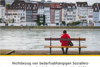 Person in roter Jacke sitzt auf einer Bank am Fluss, mit Gebäuden im Hintergrund.