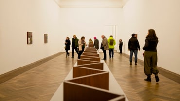 Menschen in einer Kunstgalerie mit Holzinstallation in der Mitte.