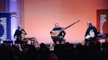 Drei Musiker auf Bühne mit Instrumenten in Konzertsaal.",