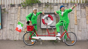 Zwei Männer in grünen Anzügen auf Tandemrad vor Steinmauer.