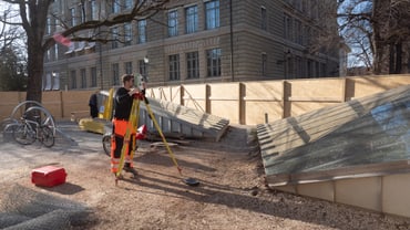 Bauarbeiter beim Vermessen neben einem Gebäude und Holzbarrieren.