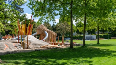 Spielplatz mit Klettergerüst und Rutsche im Park unter Bäumen.