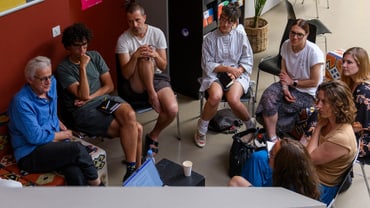 Gruppe von Menschen in einer Besprechung vor einem Regal mit Postern an einer Wand.