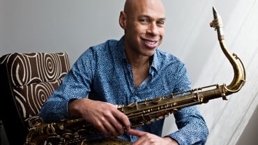 Künstler mit Saxophon auf braunen Sessel sitzend, lächelnd. 