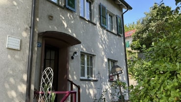 Aussenansicht eines zweistöckigen Hauses mit offenen Fenstern und umgebender Vegetation.
