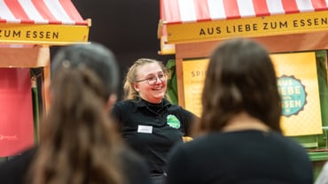 Lächelnde Frau spricht zu Publikum, Marktstände im Hintergrund.