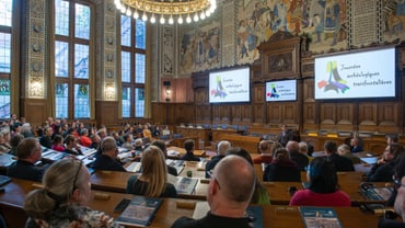 Menschen in einem Saal mit Holzwänden, zuhören einem Vortrag.
