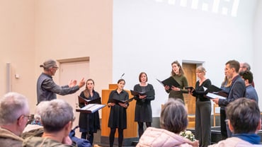 Chorprobe in einem Raum mit hellem Gewölbe, Zuschauer im Vordergrund.