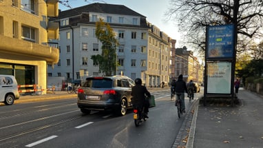 Strassenszene mit Autos und Radfahrern bei Sonnenuntergang.",