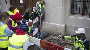 Kinder in Warnwesten beobachten einen Archäologen an einer Baustelle.