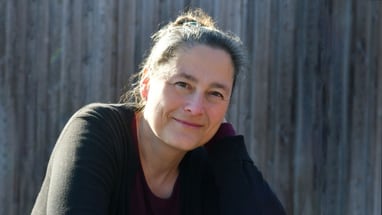 Frau mit schwarzen Pullover lächelt vor Holzwand.