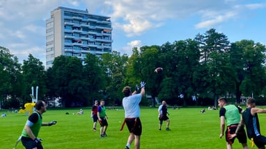 Menschen spielen Football auf einem grünen Feld mit einem Hochhaus im Hintergrund.