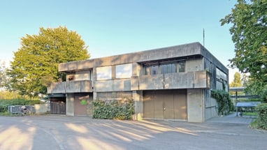 Modernes zweistöckiges Gebäude aus Beton mit Bäumen im Hintergrund.