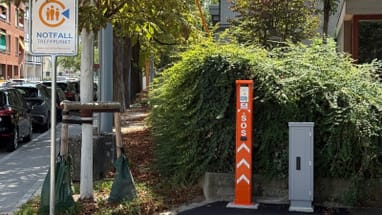 Notfalltreffpunkt-Schild und SOS-Säule nahe Gehweg mit Bäumen.