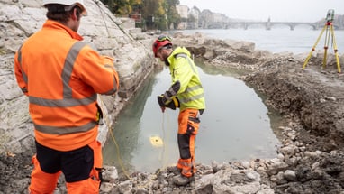 Archäologen in orangefarbener Kleidung führen Untersuchungen am Ufer des Rheins durch.