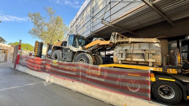 Grosser Bagger auf einem Anhänger vor einem Gerüstgebäude.