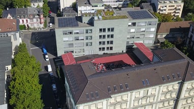 Drohnenansicht von Gebäuden mit Solarpanels und rotem Dachbereich.",