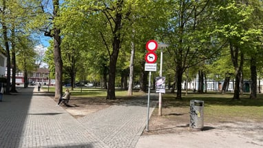 Gepflasterter Weg durch einen Park mit Bäumen und Schildern.