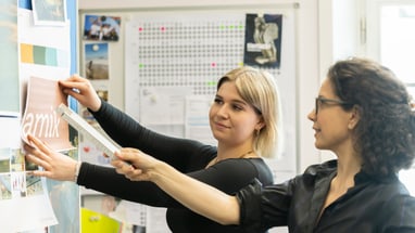 Zwei Frauen hängen Bilder an einer Pinnwand in einem Büro auf.