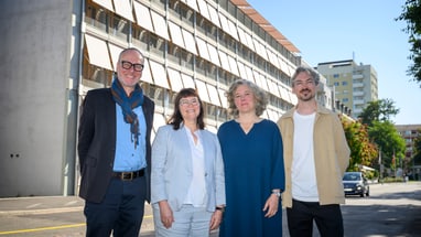Vier Personen lächeln ausserhalb eines Gebäudes in der Sonne.