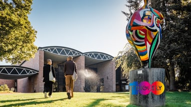 Zwei Personen gehen zum Tinguely-Museum mit bunter Skulptur davor.