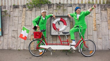 Zwei Männer in grünen Anzügen auf Tandemrad vor Steinmauer.