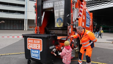 Mitarbeiter der Stadtreinigung in leuchtender Arbeitskleidung zeigt Kind die Funktion eines elektrischen Müllwagens in Basel.