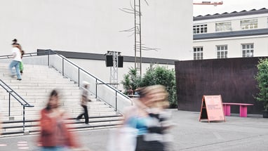 Menschen gehen draussen an einer weissen Wand vorbei.