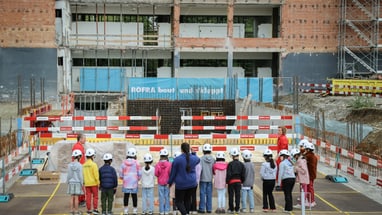 Kinder mit Helmen bei einer Baustellenbesichtigung vor einem Gebäude im Bau.