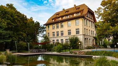 Grosses Gebäude mit Teich im Vordergrund und Bäumen im Hintergrund.