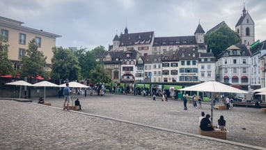 Menschen auf einem gepflasterten Platz mit historischen Gebäuden im Hintergrund.