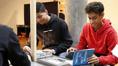 Zwei junge Männer sitzen an einem Tisch und lesen im Logbuch der Ausstellung «Mensch, du hast Recht(e)»