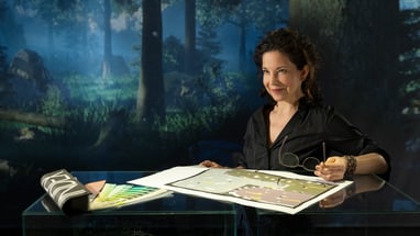 Frau sitzt am Tisch mit Zeichnungen vor einem Hintergrund mit Waldlandschaft.