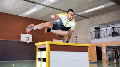 Mann springt über Hindernis in Turnhalle.