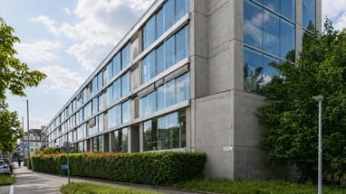 Modernes Glasgebäude an einer Strasse mit Bäumen und Büschen.