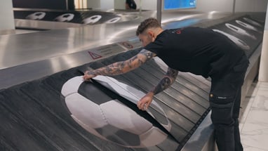 Mann prüft Förderband mit Fussballmotiv am Flughafen.