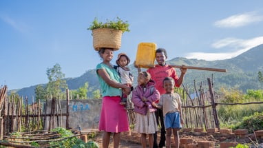 Familie in einem Garten vor einem Berg, Frau trägt Korb auf dem Kopf.