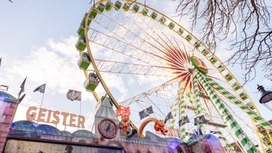 Riesenrad und Geisterbahn auf Jahrmarkt mit Baumzweigen im Vordergrund.