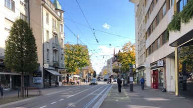 Stadtstrasse mit Geschäften und Bäumen bei Sonnenschein.",