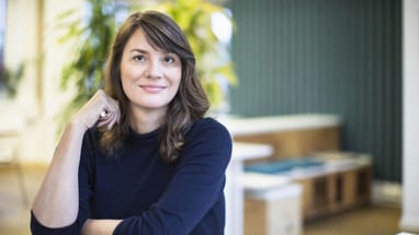 Person mit dunklem Pullover, sitzt und lächelt in einem modernen Raum.