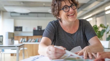 Lachende Frau mit Brille sitzt an einem Tisch.