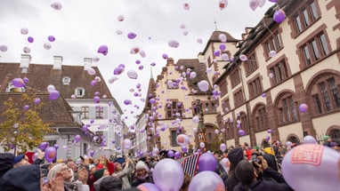 Menschenmenge mit lila Luftballons in historischer Stadtumgebung.