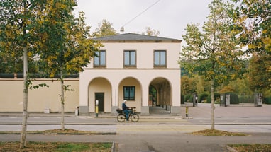 Person auf Fahrrad vor zweistöckigem Gebäude und Bäumen.