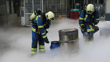 Zwei Feuerwehrleute mit Atemschutzmasken im Rauch bei Fässern.