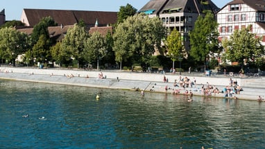 Flussufer mit Menschen und Häusern bei sonnigem Wetter.